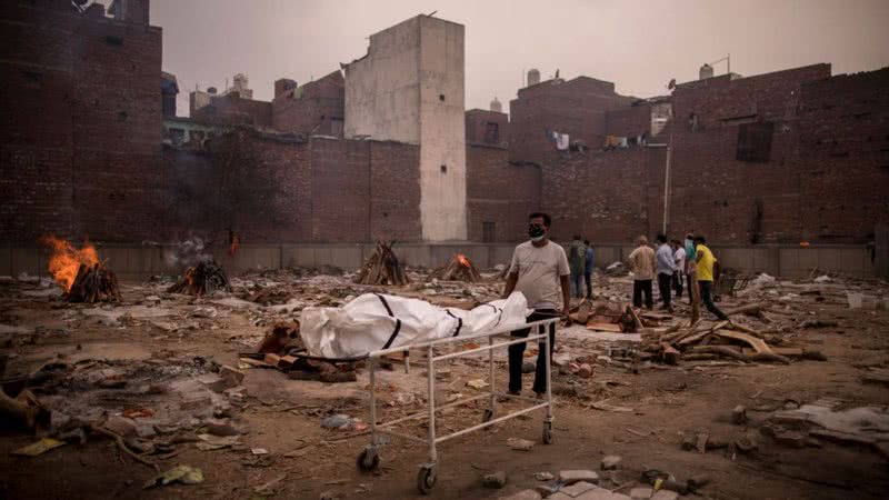 Fotografia de campo indiano onde estão sendo realizadas piras funerárias para mortos da pandemia, com homem trazendo novo cadáver - Getty Images