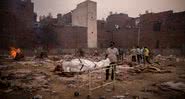 Fotografia de campo indiano onde estão sendo realizadas piras funerárias para mortos da pandemia, com homem trazendo novo cadáver - Getty Images